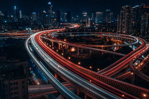 Red and white light trails from traffic curving through a city highway interchange.