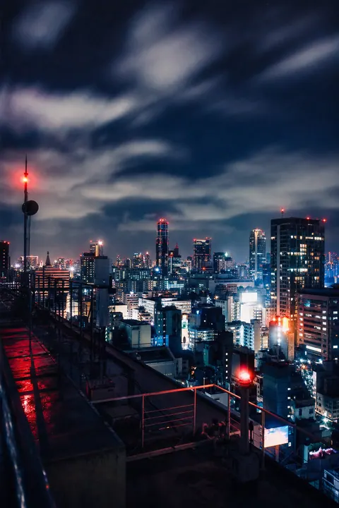 A city skyline at night under clouds streaked by a long exposure, red antenna lights on top.