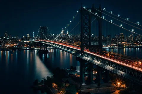 A suspension bridge at night with light trails from cars above a glassy smooth river.