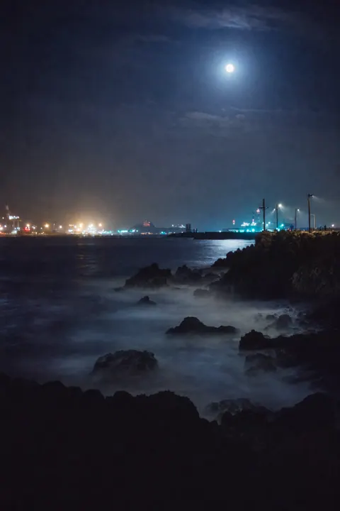 Moonlit waves blurred into mist by a long exposure, with harbour lights in the distance.