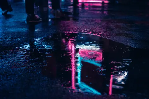 A puddle on a dark street reflecting a pink and blue neon sign, rippled by raindrops.