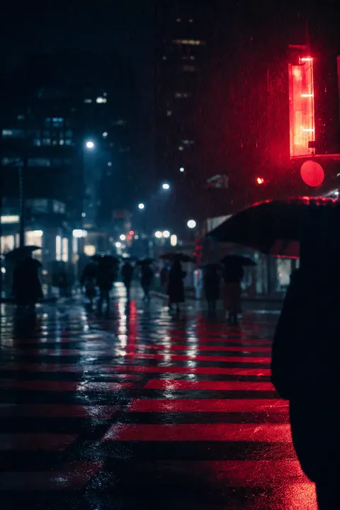 A rain-soaked zebra crossing glowing red under a neon sign, blurred umbrellas far away.