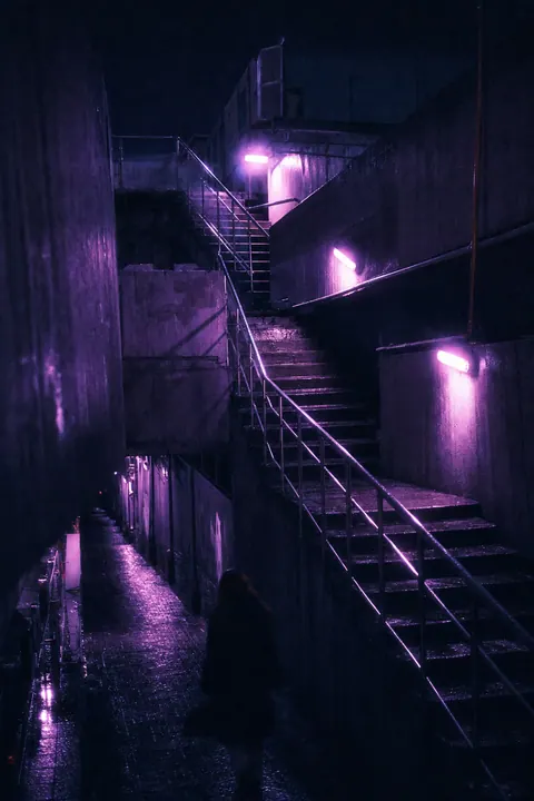 A concrete outdoor stairwell bathed in violet neon light with gleaming wet railings.