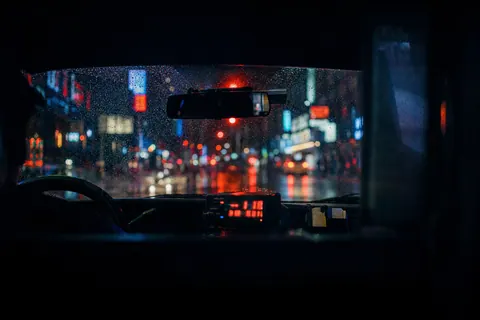 The view through a rainy taxi windshield at a red light, neon signs blurred into bokeh.