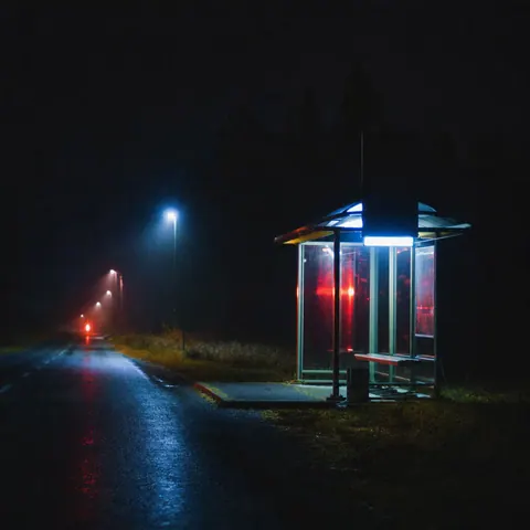 A single lit bus shelter beside an empty country road surrounded by darkness.