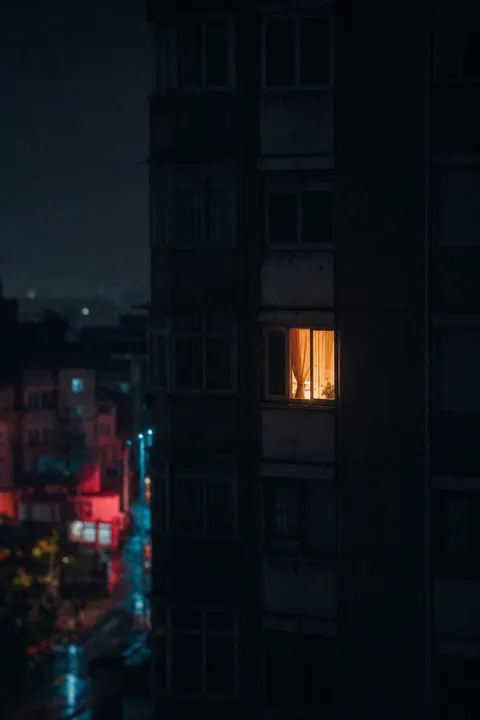 One warmly lit window in an otherwise dark apartment building at night.