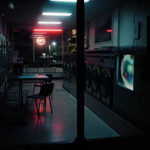 An empty fluorescent-lit laundromat at night with one washing machine running.